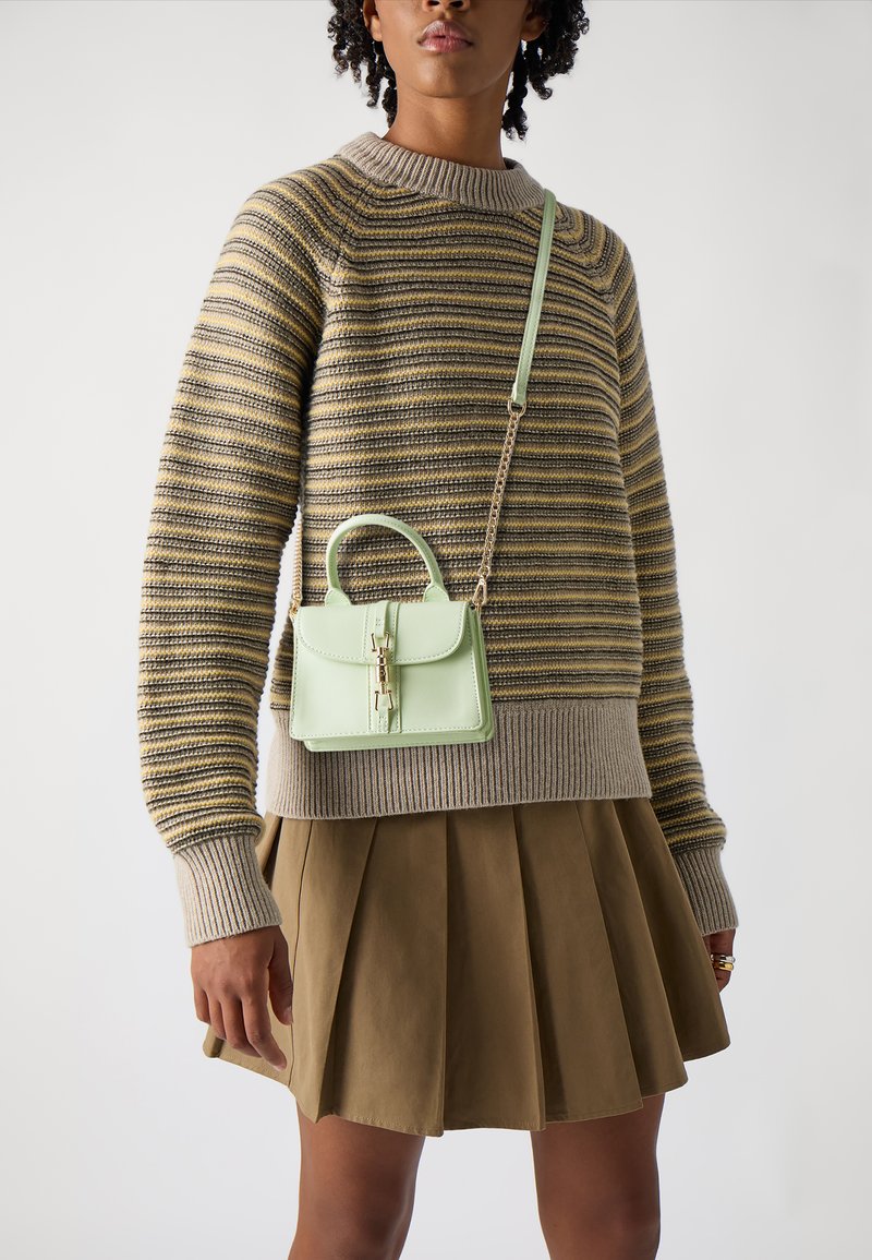Green leather handbag with a top handle and chain strap, featuring a flap closure and rectangular shape, paired with a striped sweater and pleated skirt.