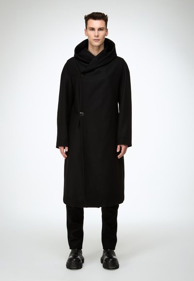 Schwarzer langer Mantel aus schwerem Stoff mit großer Kapuze, Knopfverschluss und geradem Schnitt. Trägt man über schwarzen Hosen und massiven Stiefeln.