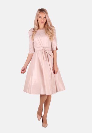 Helles rosa Satin-Kleid mit tailliertem Oberteil, schulterfreien Ärmeln, gebundenem Taillengürtel und weitem Rock, ergänzt durch elegante, hautfarbene High Heels.