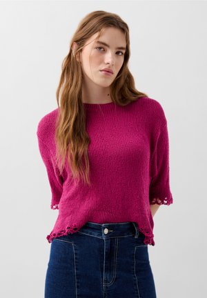 Pullover magenta lavorato a maglia con maniche corte, caratterizzato da un motivo testurizzato e orlo a scallop. Abbinato a una gonna in denim a vita alta.