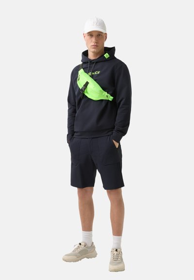 Black hoodie with "PRE-ICE" logo, neon green sling bag, navy shorts, and beige sneakers with white socks, on a neutral background.