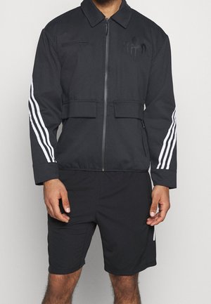 Mann trägt eine schwarze Jacke mit Reißverschluss und weißen Streifen an den Ärmeln sowie passende schwarze Shorts vor einfarbigem Hintergrund.
