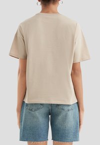 Beige short-sleeve t-shirt with a relaxed fit and ribbed crew neckline, shown from the back. Material appears soft with a smooth texture.