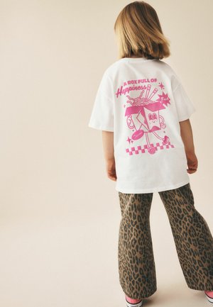 Barn med skulderlangt hår bærer en hvid oversized T-shirt med en pink tegneserie nudelæske og tekst, leopardmønstrede bukser og pink sokker.