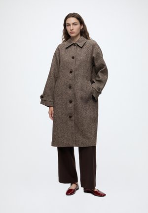 Femme debout portant un long manteau marron boutonné, un pantalon large foncé et des chaussures plates rouges sur un fond clair uni.