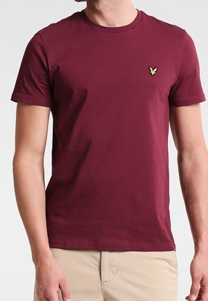 Mand iført en ensfarvet bordeaux kortærmet t-shirt med et lille gult logo på venstre bryst, kombineret med beige bukser.