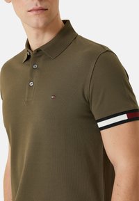 Camisa polo de color verde oliva con tejido texturizado, dos botones y un puño a rayas en contraste que presenta acentos en rojo, blanco y negro.