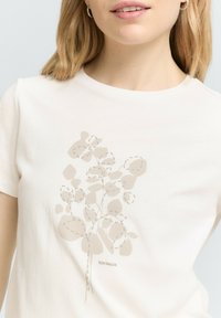T-shirt beige clair avec un design botanique en beige plus foncé, avec des contours en pointillés et un logo en bas. Manches courtes et col rond.
