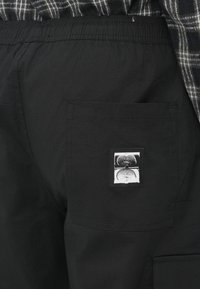 Pantalon cargo noir avec ceinture élastique, poche latérale et étiquette carrée avec un motif graphique sur la poche arrière.