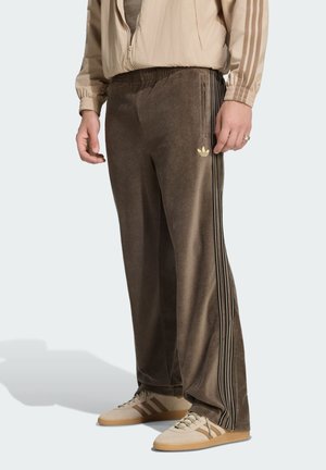 Uomo che indossa pantaloni sportivi marroni Adidas con tre strisce laterali, scarpe beige e giacca beige con cerniera, in piedi su sfondo semplice.