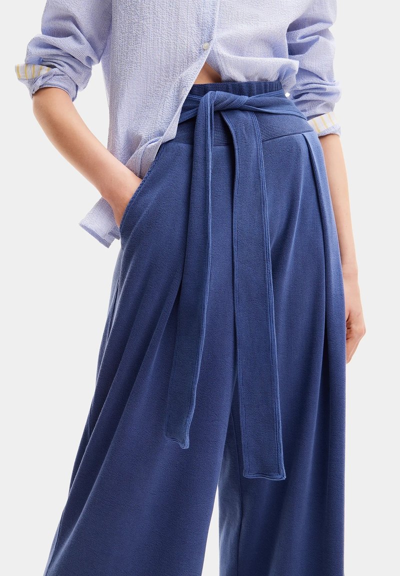 Pantaloni a gamba larga blu in tessuto testurizzato, con dettaglio in vita legato e tasche laterali. Abbinati a una camicia a righe blu chiaro.