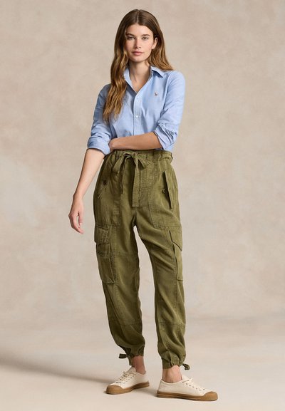 Naine, kellel on pikad juuksed, seljas hele sinine nööbitav särk, oliivrohelised lahtised püksid lipsuga vööl ning beežid spordijalatsid, seisab ühe käe üle rinna risti.