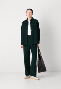 Camicetta di velluto a coste verde scuro e pantaloni abbinati con una t-shirt bianca sotto. Scarpe argentate e una borsa a tracolla nera. Design classico con bottoni e tasca.