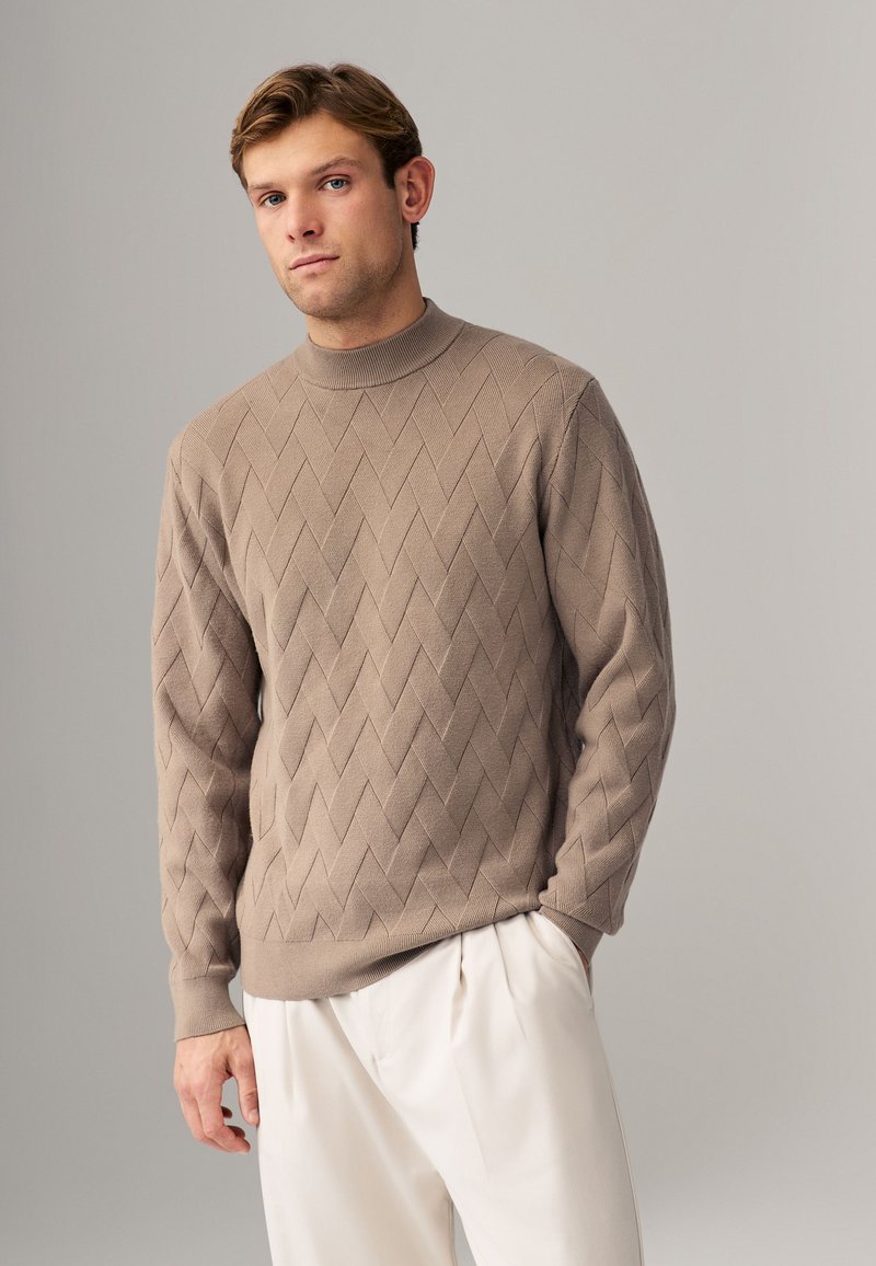 Beige Strickpullover mit raised Diamantmuster, hohem Kragen und gerippten Bündchen. Kombiniert mit hellen, plissierten Hosen.