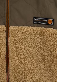 Pihepárnázott zöld felső anyag, barna bolyhos alsó rész, látható narancssárga cipzár, valamint fekete címke fehér szöveggel és földgömb logóval.