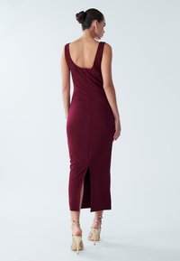 Burgundy fitted dress with a scoop back, knee-length slit, and smooth fabric. Worn with strappy beige heels. Back view visible.