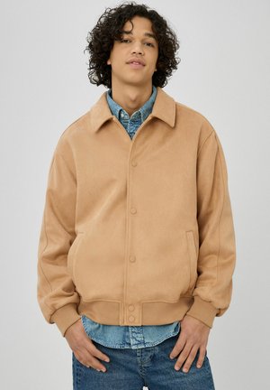 Jeune personne aux cheveux bouclés portant une veste beige boutonnée par-dessus une chemise en jean bleue et un jean, debout devant un fond uni.