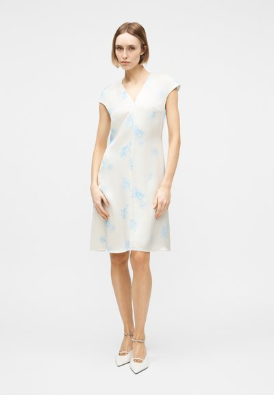 Γυναίκα που φοράει λευκό φόρεμα μήκους μέχρι το γόνατο με ανοιχτό μπλε λουλουδάτα σχέδια και λευκά στιλέτο παπούτσια με λουράκια στον αστράγαλο, στέκεται μπροστά σε λευκό φόντο.