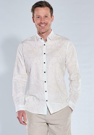 Lächelnder Mann mit kurzen braunen Haaren, der ein helles gemustertes Hemd und beigen Hosen trägt, stehend vor einem schlichten Hintergrund.