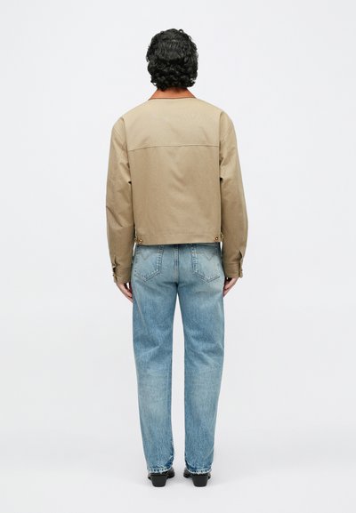 Vyras su tamsiais garbanotais plaukais, dėvintis rudą trumpą švarkelį ir šviesiai mėlynas džinsus, stovintis nugara į priekį ant balto fono.