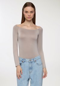 Body na długi rękaw w jasnym taupe, fason typu off-shoulder z gładką teksturą. Połączony z jeansami w stylu high-waisted w wyblakłym niebieskim odcieniu.