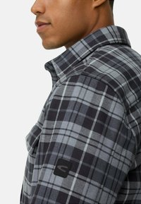 Chemise en flanelle à carreaux dans des tons de noir et de gris. Elle possède un col, une poche visible et une étiquette logo noire sur la manche.