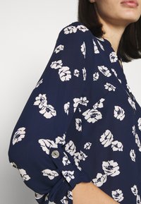 Námořnická blůza vyrobená z lehkého materiálu s bílým květinovým vzorem. Obsahuje knoflíkové detaily na ohrnutých rukávech a má klasický design blůzy.