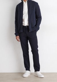 Marinblå zip-up jacka med hög krage, i kombination med mörk marinblå byxor. Vit skjorta under och vita sneakers fulländar looken.