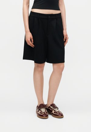 Persoon draagt zwarte op maat gemaakte shorts tot op de knieën, een zwarte cropped top en bruine leren sneakers met witte veters.