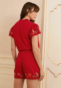 Tutina rossa con maniche corti a sbuffo, bordi ondulati e ricami floreali che presentano uccelli e piante sulle maniche e sull'orlo. Tessuto morbido.