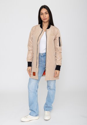 Femme aux longs cheveux foncés portant une veste bomber beige avec des poignets noirs, un jean bleu clair, des baskets blanches et un haut blanc, debout devant un fond uni.