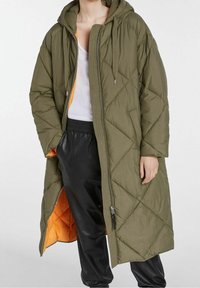 Vadderad lång kappa i olivgrönt med huva; har ett orange foder, metallisk dragkedja och diagonala sömnadsmönster. Svarta byxor syns.