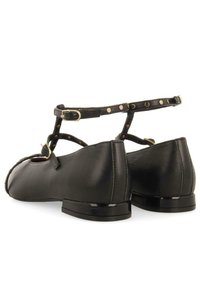 Zapatos planos de cuero negro con punta redonda, tira en el tobillo y detalles de tachuelas. Tacón bajo y brillante en negro. Textura suave y diseño clásico.