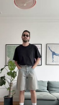 Fekete, oversize póló, világosszürke farmer rövidnadrág, fekete napszemüveg és fehér zokni. A háttérben növények és keretezett műalkotás látható.