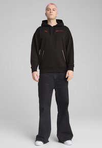 Hoodie noir avec poche kangourou, arborant le logo rouge de la Formula 1 et la marque Puma. Porté avec un jean noir évasé et des baskets blanches.