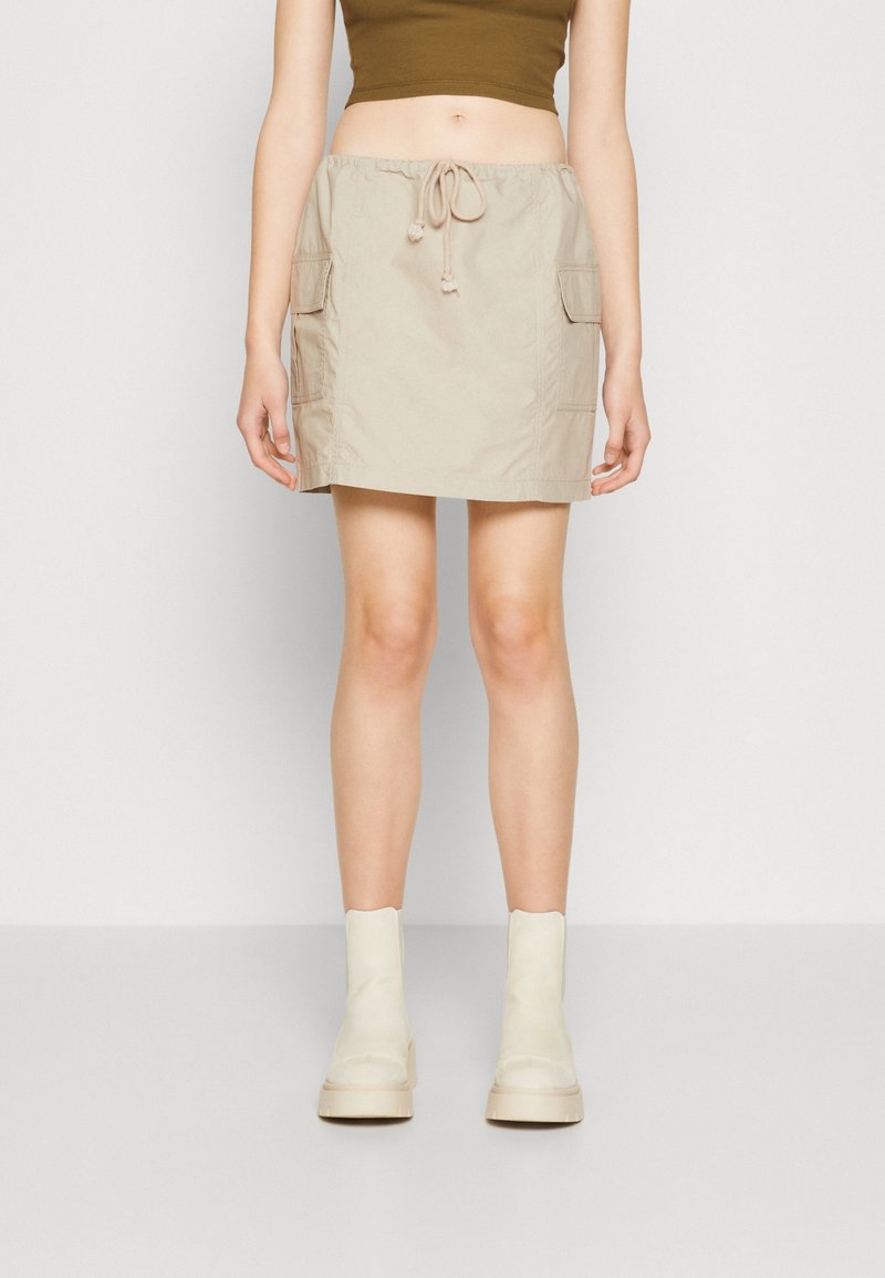Femme portant une mini-jupe cargo beige avec taille à cordon, un crop top olive et des bottines blanches, debout devant un fond uni.