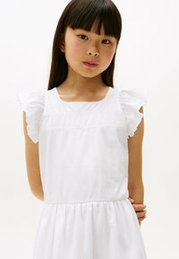 Vestido de algodón blanco con tejido texturizado, que presenta tirantes de hombro con volantes y una cintura fruncida. Diseño simple y clásico.