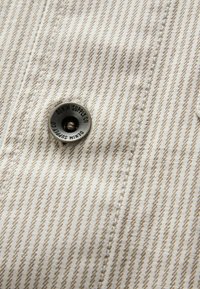 Bouton étiqueté "Denim Supply Co" sur un tissu rayé beige et blanc avec des détails visibles de couture.