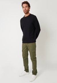 Marine kabelgebreide trui, olijfgroene cargobroek en witte sneakers. De outfit heeft een gestructureerde stof en praktische zakken op de broek.