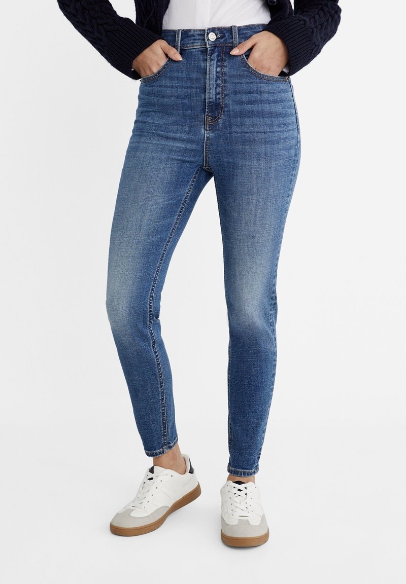 Schlanke blaue Denim-Jeans mit hoher Taille, vorderen Taschen und leichtem Verblassen. Kombiniert mit weißen Sneakers mit hellgrauen Akzenten.