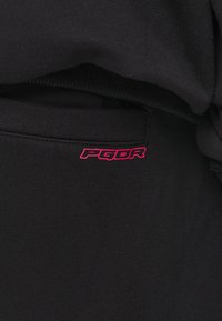 Svart tyg med en slät yta som har en rosa broderad "PGDR"-logotyp på fickan. Enkel, modern design med en avslappnad stil.