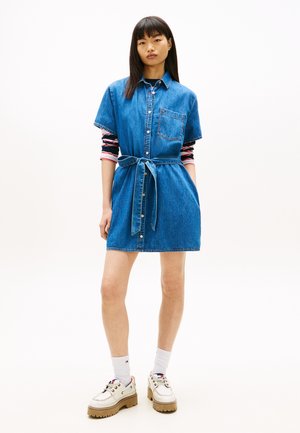 Donna che indossa un vestito di jeans blu a maniche corte con bottoni frontali, cintura annodata, maglia a righe a maniche lunghe sotto, calzini bianchi e scarpe chunky.