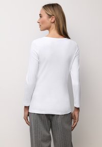 Wit katoenen top met lange mouwen en een ronde halslijn. Gladde textuur, aansluitend ontwerp, bekeken van de achterkant, gecombineerd met grijze gestreepte broek.