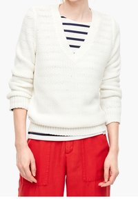 Pull en maille blanche avec col en V et design texturé, superposé à une chemise rayée bleu marine et blanc, associé à un pantalon rouge.