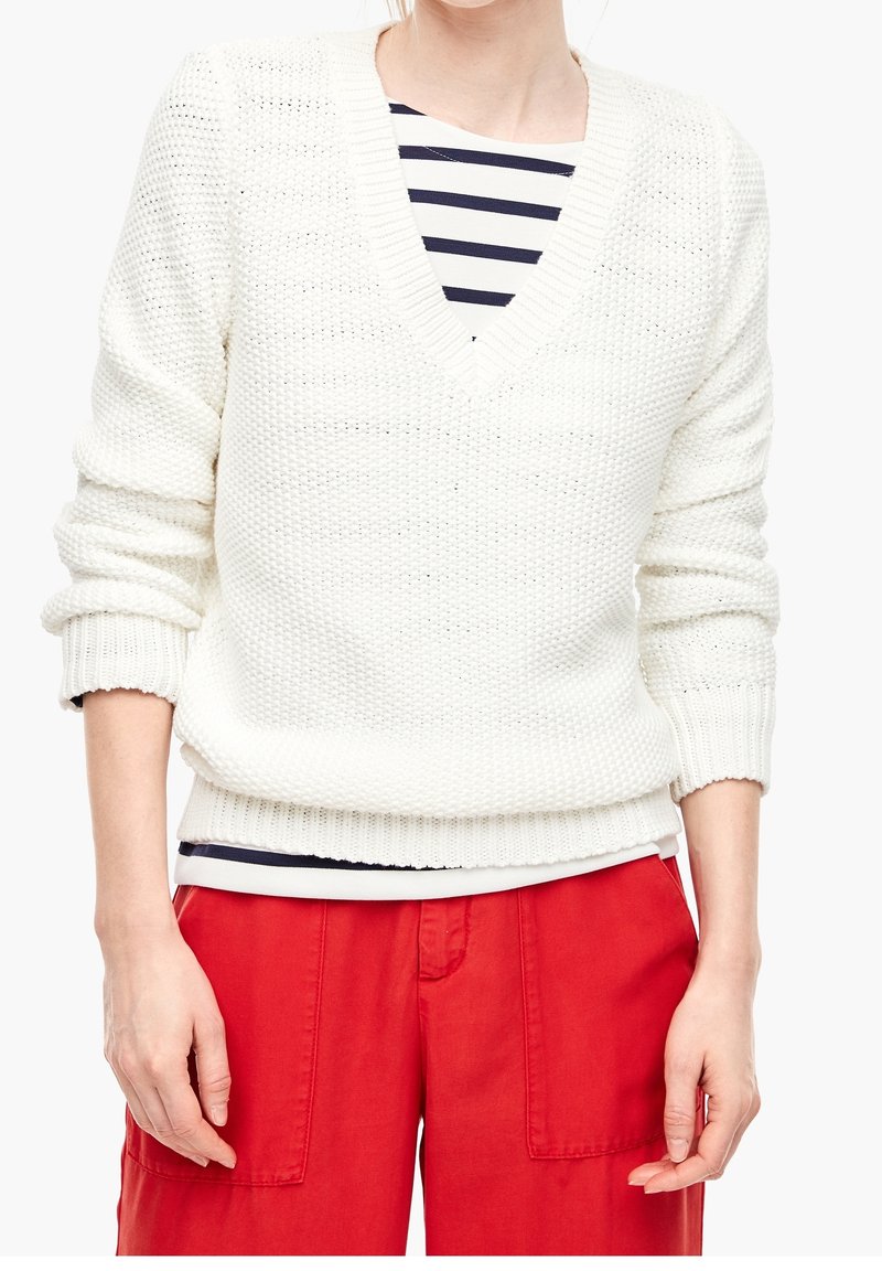 Pull en maille blanche avec col en V et design texturé, superposé à une chemise rayée bleu marine et blanc, associé à un pantalon rouge.