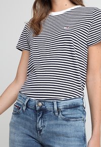 Camiseta a rayas en tejido azul marino y blanco, con escote redondo, mangas cortas y pequeño detalle de logo en el pecho, combinada con jeans de mezclilla claros.