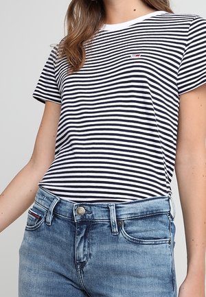 Camiseta a rayas en tejido azul marino y blanco, con escote redondo, mangas cortas y pequeño detalle de logo en el pecho, combinada con jeans de mezclilla claros.