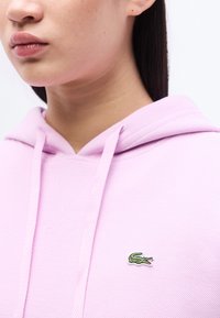 Κοντινό πλάνο ενός ατόμου που φοράει ροζ ανοιχτό φούτερ με μικρό πράσινο λογότυπο κροκόδειλου στο στήθος, με ίσια σκούρα μαλλιά να φαίνονται πάνω από τον ώμο.