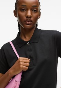 Fekete poloing gallérral, gombos nyakkal és steppelt mintával. A modell egy világos rózsaszín táskát tart, aminek pántja van.