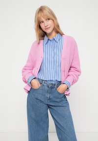 Cardigan rosa tricotado com botões, usado sobre uma camisa riscada azul clara. Modelo está com jeans azuis de cintura alta, com as mãos nos bolsos.